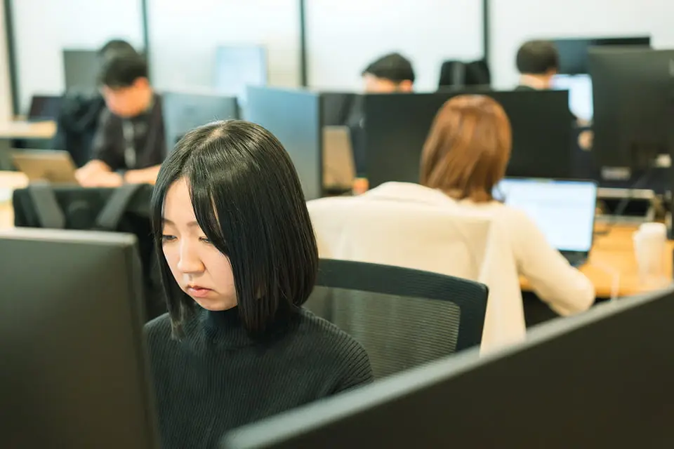 オフィスでパソコン作業に集中する女性と働く同僚たちの様子