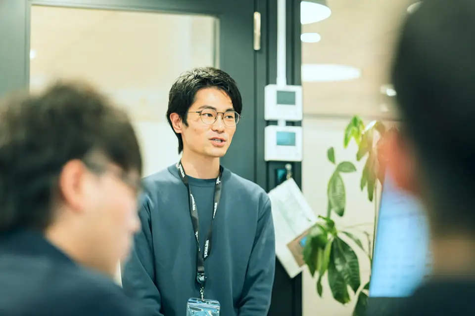 オフィスで説明を行う社員と、その話を聞く複数の同僚たちを捉えたシーン